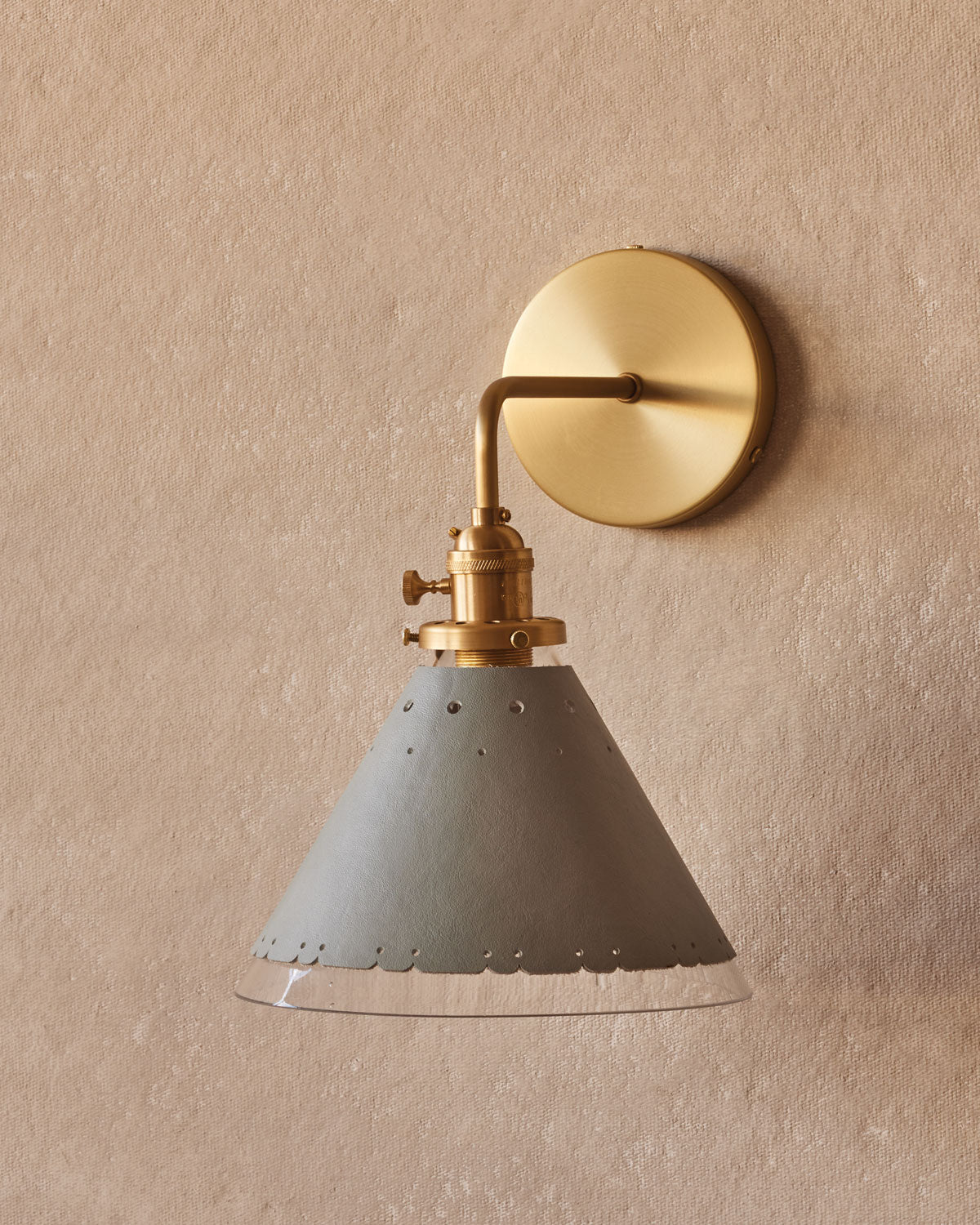 A wall-mounted Stella Sconce with a hand-blown glass shade and a perforated leather sleeve in a neutral color, with a circular backplate. #leather-finish_cloud blue #wiring_hardwired